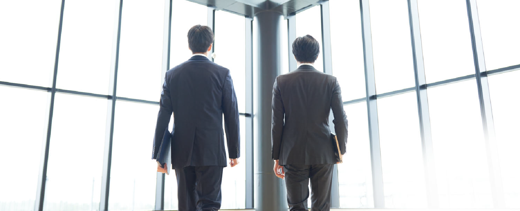 大きな窓のあるオフィスビル内を、スーツ姿の2人のビジネスマンが並んで歩いている後ろ姿